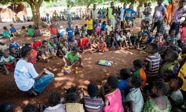 Molti bambini siedono in cerchio con un collaboratore SOS e guardano un bambino al centro.